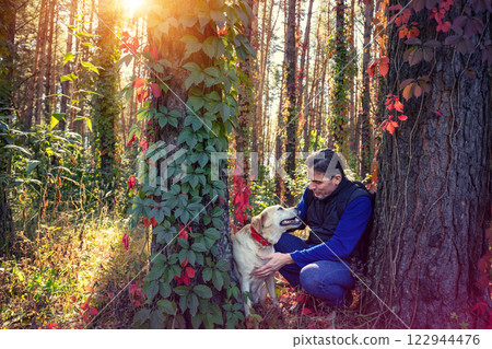 Happy man with Labrador retriever dog walking in the pine forest in autumn. Man and dog sitting among two trees 122944476