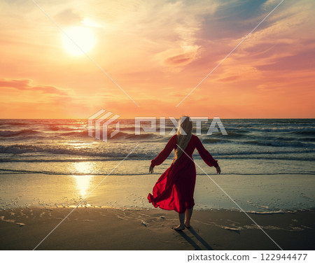 Seascape during orange sunrise with beautiful sky. Woman on the beach. Young happy woman in a long red fluttering dress walks on the seashore Seascape during orange sunrise with beautiful sky. Woman on the beach. Young happy woman in a long red fluttering dress walks on the seashore 122944477