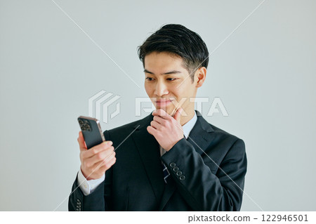 A young man in his twenties holding a smartphone with his hand on his chin 122946501