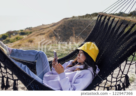 A young attractive Asian woman holds a mobile phone and lies in a hammock. A charming Korean woman in casual clothes uses a smartphone and relaxes in nature. 122947366