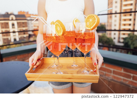Close-up of young girl holding wooden tray with four glasses of aperol. Delicious citrus alcohol cocktails with slice of orange. Close-up of young girl holding wooden tray with four glasses of aperol. Delicious citrus alcohol cocktails with slice of orange. 122947372
