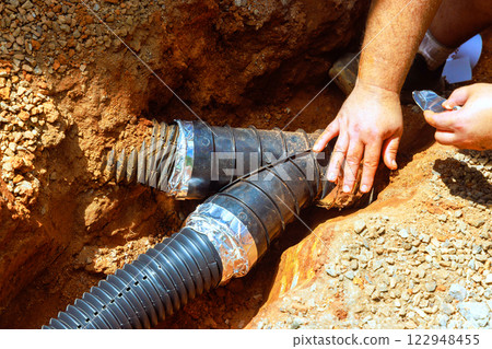 Worker connects black plastic drainage pipes in trench at construction site, ensuring proper installation amidst dirt. 122948455