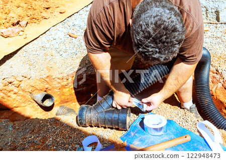 Worker is busy connecting pipes, installing drainage systems in newly excavated trench outdoors. 122948473