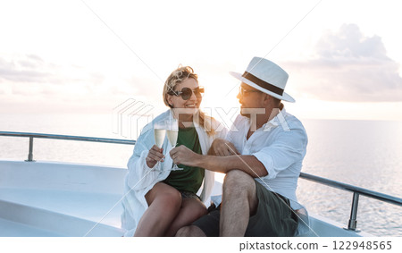 A young married couple meets the sunset at the stern of the ship. A smiling woman in sunglasses clinks glasses of champagne with her husband. 122948565
