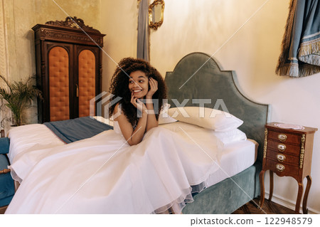 A young cute African woman in a white dress on a hotel bed. Retro stylish interior. A pretty black girl in a hotel room. A young cute African woman in a white dress on a hotel bed. Retro stylish interior. A pretty black girl in a hotel room. 122948579