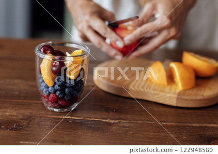 Close-up of a glass bowl with fruits and berries and a female hands. A young woman is slicing a fresh fruit salad. Close-up of a glass bowl with fruits and berries and a female hands. A young woman is slicing a fresh fruit salad. 122948580