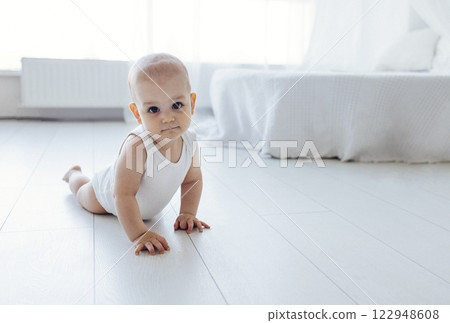 Sweet baby crawls on the floor of the bedroom. Cute little child in white bodysuit looks at the camera. 122948608