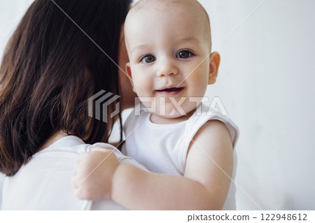 Pretty infant in a white bodysuit with his mom on a light background. Motherhood concept. Pretty infant in a white bodysuit with his mom on a light background. Motherhood concept. 122948612