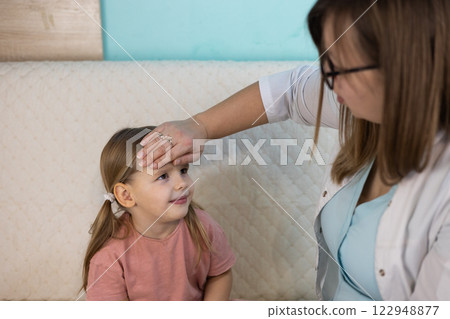 A female pediatrician measures the temperature of a sick little girl at home, placing her hand on her forehead. The doctor provides individual medical care to the child in a comfortable environment. 122948877