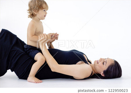 Young pregnant woman is lying on floor and playing with her son. Adorable mom and her little boy are having fun together. White isolated background. 122950023