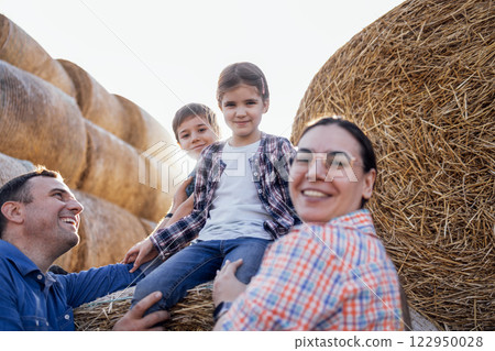 A young happy family of farmers in casual clothes. A smiling little boy and a girl are sitting on a haystack on a farm. 122950028