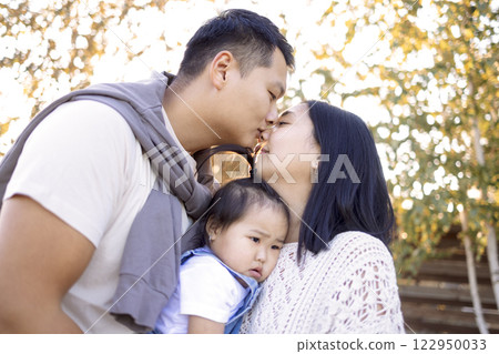 An Asian couple in love is kissing outdoors and holding their kid. Cute affectionate Korean parents in casual clothes hug their little daughter. A happy family. An Asian couple in love is kissing outdoors and holding their kid. Cute affectionate Korean parents in casual clothes hug their little daughter. A happy family. 122950033