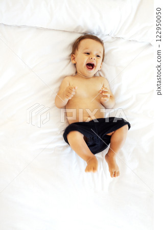 The cute baby is laughing and lying on a white sheet on the couch. A little boy is smiling on the bed at home. 122950050