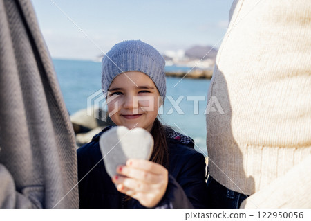 Cute little girl stands between her parents and shows a heart-shaped stone. A sweet baby in a knitted hat and blue jacket on a pebble beach with her parents. Cute little girl stands between her parents and shows a heart-shaped stone. A sweet baby in a knitted hat and blue jacket on a pebble beach with her parents. 122950056