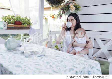 Young beautiful mother drinks tea and holds cute baby in white bodysuit in her arms. Young beautiful mother drinks tea and holds cute baby in white bodysuit in her arms. 122950062