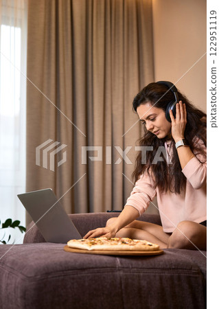 Caucasian woman working on laptop computer and eating pizza on sofa in living room Caucasian woman working on laptop computer and eating pizza on sofa in living room 122951119