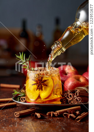 Apple cider is poured from a bottle into a glass with apple slices, anise, and a rosemary branch. 122951677