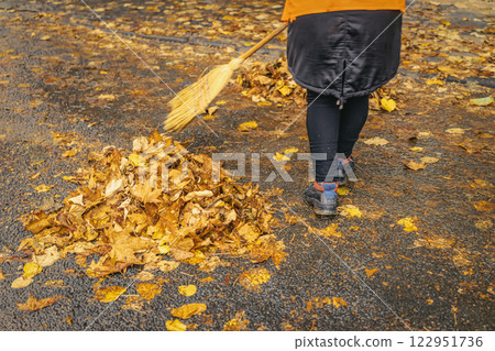 Worker in orange jacket sweeping fallen autumn leaves on wet pavement. Seasonal cleanup, urban maintenance in rainy weather 122951736