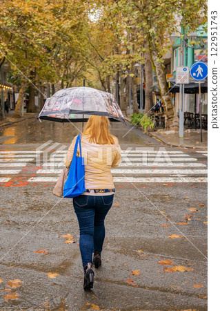 Woman in beige jacket walking across wet crosswalk with transparent umbrella. Autumn rain and golden leaves create nostalgic urban mood Woman in beige jacket walking across wet crosswalk with transparent umbrella. Autumn rain and golden leaves create nostalgic urban mood 122951743