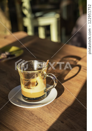 Glass cup with hot golden turmeric latte on wooden table in sunlight. Warm autumn mood, cozy coffee shop atmosphere Glass cup with hot golden turmeric latte on wooden table in sunlight. Warm autumn mood, cozy coffee shop atmosphere 122951752