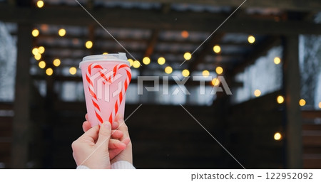 Winter pavilion glows behind pink cup and candy cane heart, where lights glitter against cool backdrop, letting lights enhance holiday mood, as lights underscore cheerful romance in frosty air. 122952092