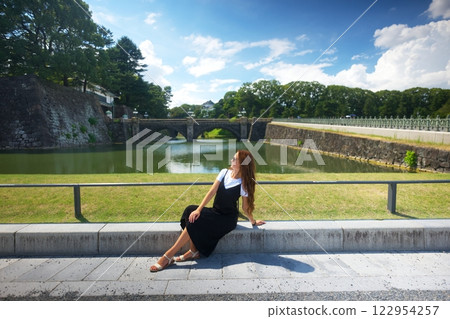 Woman Sitting by Moat with Bridge and Greenery in Tokyo on a Sunny Day Woman Sitting by Moat with Bridge and Greenery in Tokyo on a Sunny Day 122954257