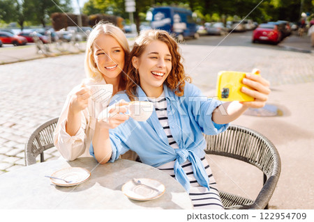 Young women taking selfie while sitting on terrace on sunny day. Girlfriends drink coffee with phone 122954709
