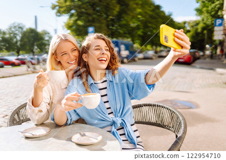 Young women taking selfie while sitting on terrace on sunny day. Girlfriends drink coffee with phone 122954710
