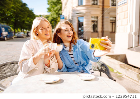 Young women taking selfie while sitting on terrace on sunny day. Girlfriends drink coffee with phone 122954711