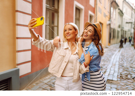 Happy female tourists in casual clothes walk along streets of Europe, take selfies. Active lifestyle Happy female tourists in casual clothes walk along streets of Europe, take selfies. Active lifestyle 122954714