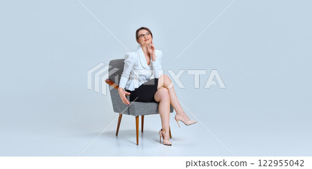 Young woman sitting on chair with legs crossed, resting hand on chin, looking thoughtful against light blue studio background. 122955042