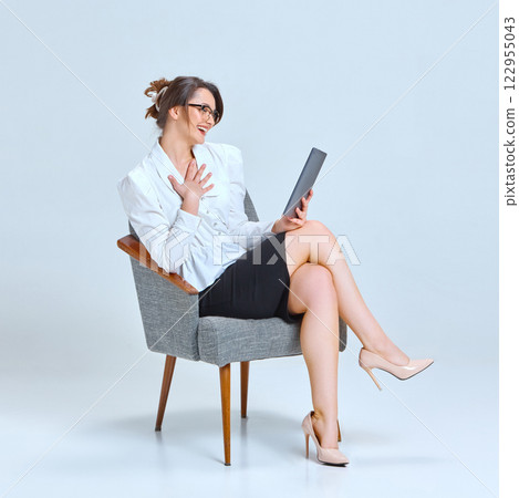 Young woman sitting on chair, wearing glasses, holding tablet, smiling warmly, engaged in conversation against light blue studio background. 122955043