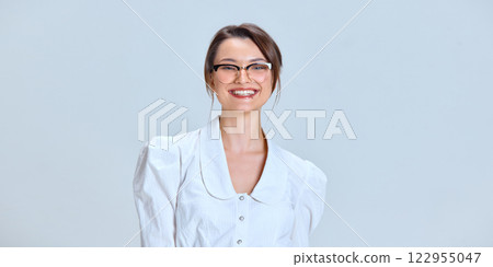 Young woman with bright smile wearing glasses, standing in white blouse, looking directly, expressing confidence against white studio background. 122955047