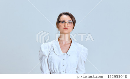 Serious young woman, in white blouse and glasses, maintaining professional posture against light blue studio background. 122955048