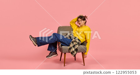 Relaxed young woman, sitting on chair, pouting with playful expression, wearing yellow hoodie against pink studio background. 122955053
