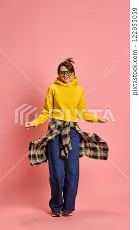 Young woman, with carefree expression, twirling checkered shirt while moving, wearing yellow hoodie, against pink studio background. Young woman, with carefree expression, twirling checkered shirt while moving, wearing yellow hoodie, against pink studio background. 122955059