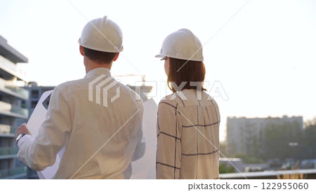 Man and woman architects constructor engineers wearing safety glasses and hardhats, are examining blueprints at a construction site during a sunset, back view. Architecture concept 122955060