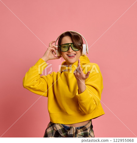 Young woman, with happy expression, singing while adjusting headphones, wearing yellow hoodie and checkered shirt, against pink studio background. 122955068