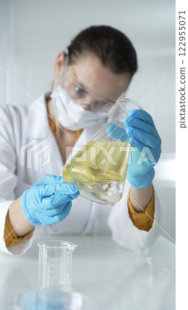 Woman scientist, wearing a lab coat, blue protective gloves, is holding an Erlenmeyer flask with yellow oily liquid in laboratory setting, portrait and vertical view. Medicine, science concept 122955071