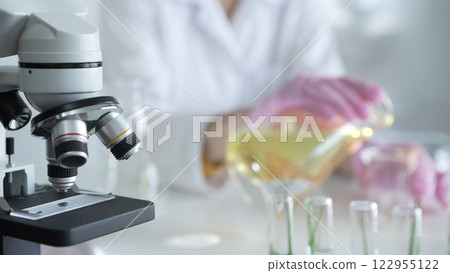 A scientist, wearing a lab coat and pink protective gloves, is pouring a yellow oily liquid from one beaker to another near microscope in laboratory, close up. Medicine and science 122955122