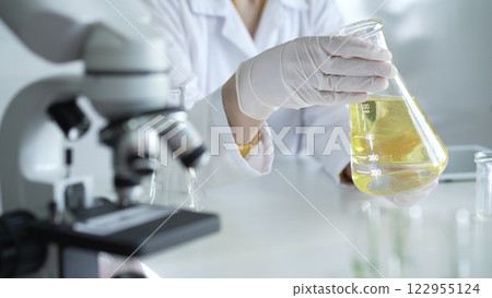 Microscope is near a scientist, wearing a lab coat and white protective gloves, and holding beaker with a yellow oily liquid in laboratory setting, close up. Medicine and science concept Microscope is near a scientist, wearing a lab coat and white protective gloves, and holding beaker with a yellow oily liquid in laboratory setting, close up. Medicine and science concept 122955124