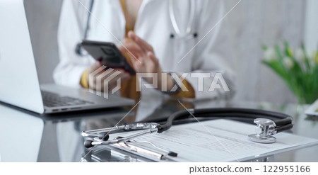 Close-up of doctor's desk with stethoscope and clipboard. Physician is using a tablet at the background. Medicine and health care 122955166