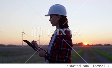 Woman engineer is taking notes on a clipboard on a field with wind turbines, as the sun sets in evening. Clean energy and engineering 122955197