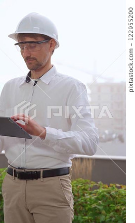 Male construction engineer or architect, wearing a white shirt and helmet, is using a digital tablet while inspecting a building site in foggy weather, front view. Architecture concept 122955200