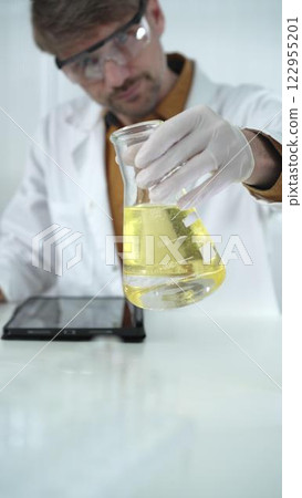 Man scientist wearing white protective gloves, and glasses is holding a yellow chemical solution inside an Erlenmeyer flask in a laboratory, vertical portrait 122955201