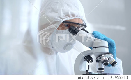 Scientist in ppe analyzing samples using a microscope in a laboratory setting. Close-up of a focused researcher in a lab coat mask and protective gear 122955213