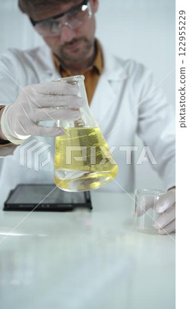 Man scientist wearing white protective gloves, and glasses is holding a yellow chemical solution inside an Erlenmeyer flask in a laboratory, vertical portrait. Science and medicine 122955229