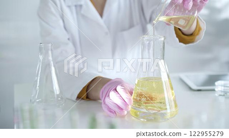 A scientist, wearing a lab coat and pink protective gloves, is pouring a yellow oily liquid from one beaker to another in laboratory, close up. Medicine and science 122955279