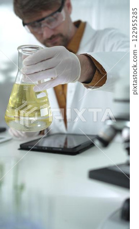 Man scientist wearing white protective gloves, and glasses is holding a yellow chemical solution inside an Erlenmeyer flask in a laboratory, vertical portrait Man scientist wearing white protective gloves, and glasses is holding a yellow chemical solution inside an Erlenmeyer flask in a laboratory, vertical portrait 122955285