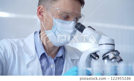 Man scientist with protective glasses, mask and gloves is using microscope in laboratory, portrait of researcher. Microbiology science concept 122955286